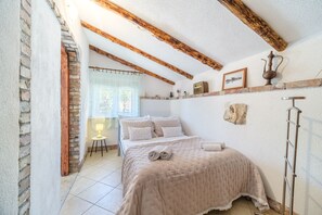 A bedroom with a bed, wooden beams, a window with curtains, a small table with a lamp, and a radiator.
