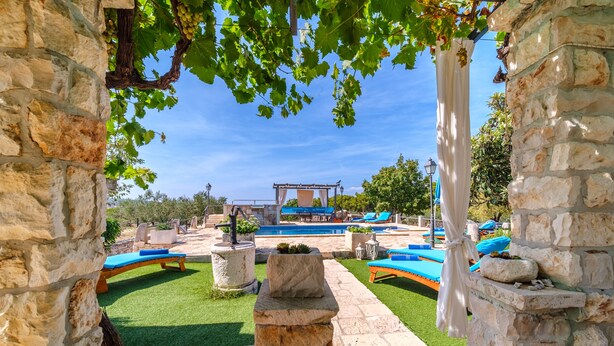 Une arche en pierre encadre un espace piscine agrémenté de chaises longues bleues et d’une pergola.