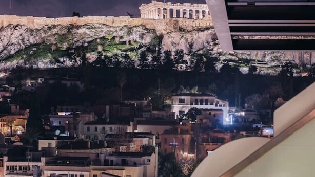 Un balcón con barandilla de vidrio que ofrece vistas a un paisaje urbano nocturno, incluyendo un edificio histórico y edificios residenciales.