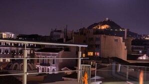 Una piscina en la azotea con cerramiento de vidrio y una chimenea visible, con vistas a un paisaje urbano con edificios iluminados y una colina al fondo.