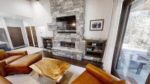 A modern living room with a stone fireplace, leather sofas, a wooden coffee table, and a large window overlooking a deck.