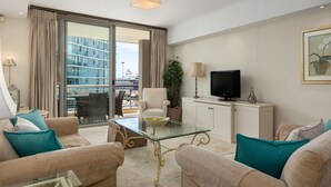 A modern living room with a glass coffee table, beige sofas, a flat-screen TV, and a view of a building through large windows.
