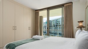 A hotel room with a large bed, a green throw blanket, beige curtains, and a view of a modern building through the window.