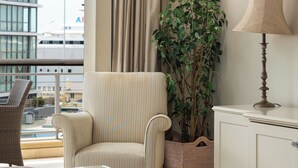 A hotel room with a view of a building, a striped armchair, a glass table with a bowl, a lamp, and a potted plant.