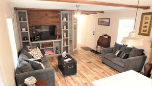 Una sala de estar acogedora con un sofá, una mesa de centro y un televisor. Hay estantes con adornos y una puerta al fondo.