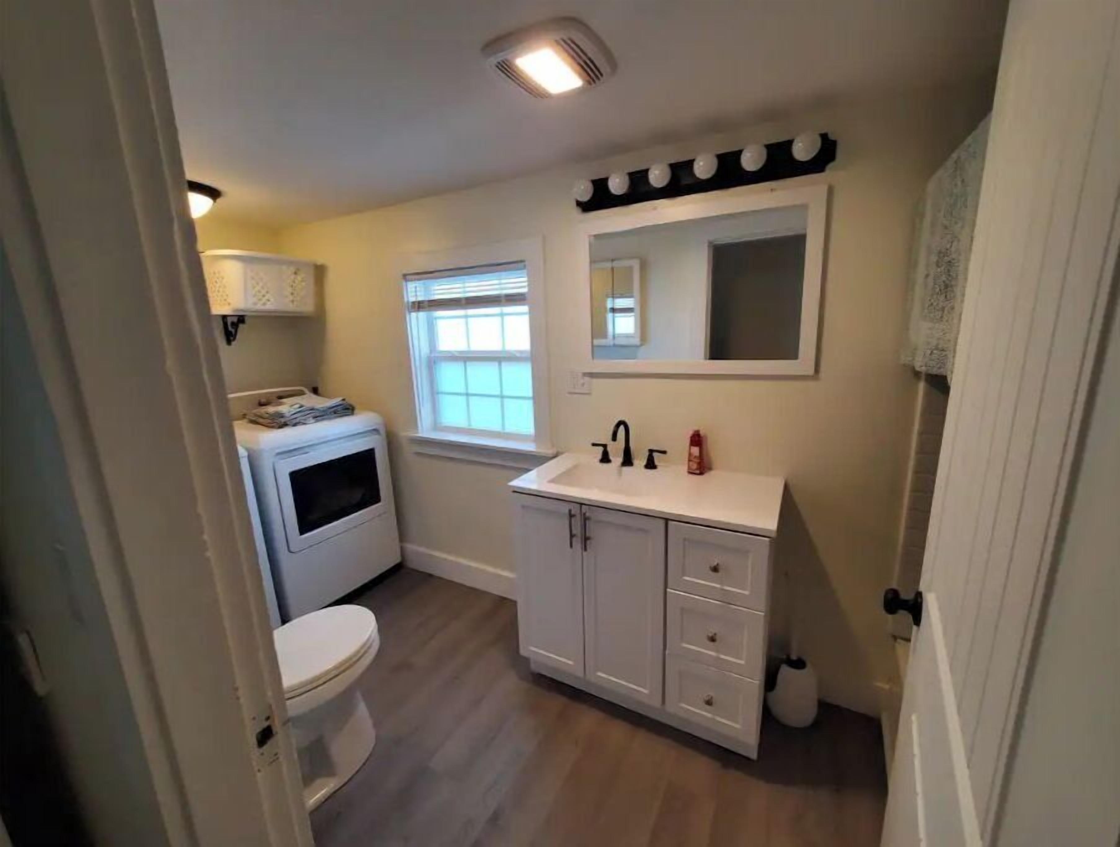 A compact bathroom with a toilet, a washing machine, a sink with a mirror, and a window.