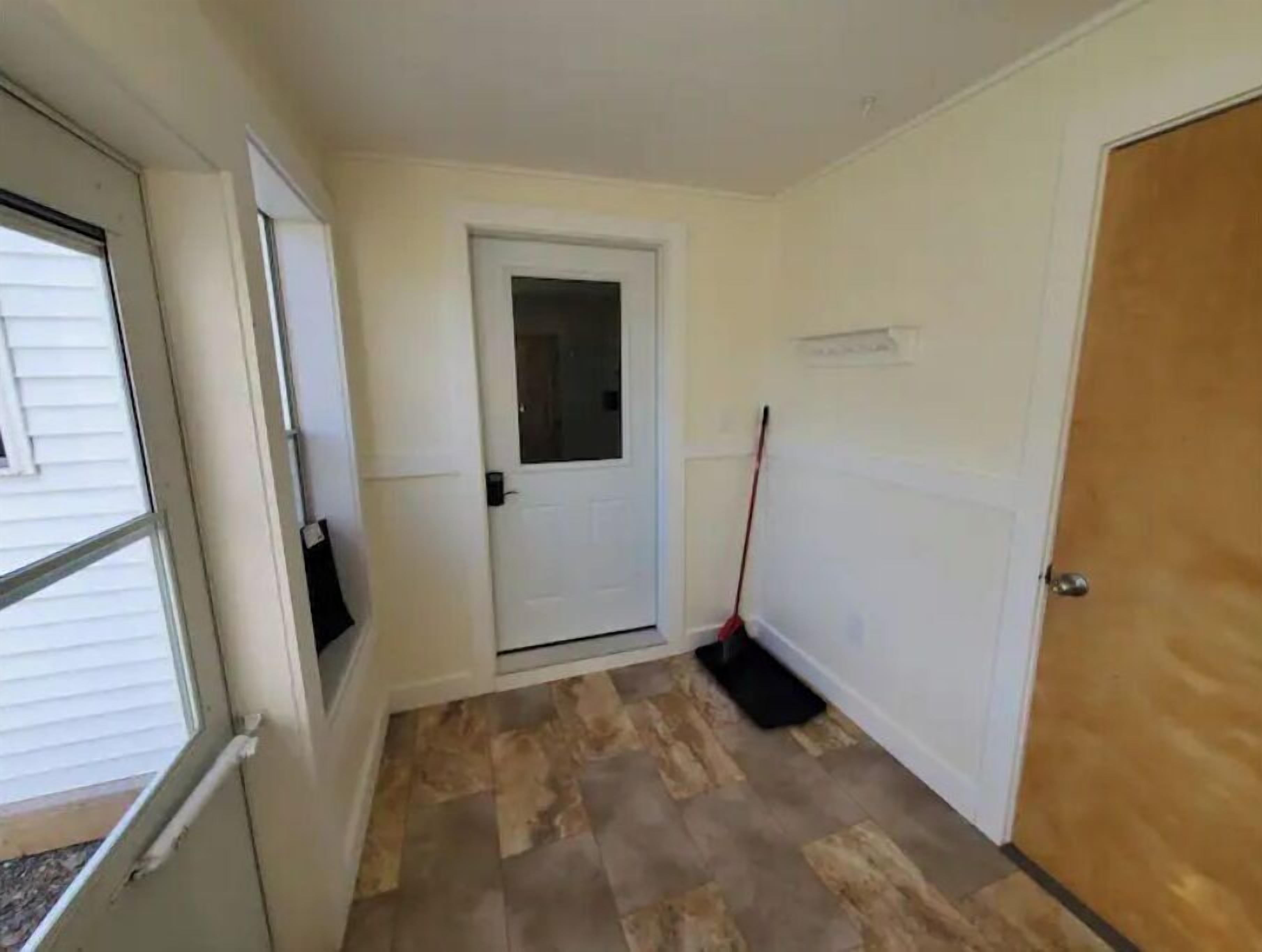 Un couloir avec une porte blanche, un balai appuyé contre le mur et une fenêtre avec des stores.
