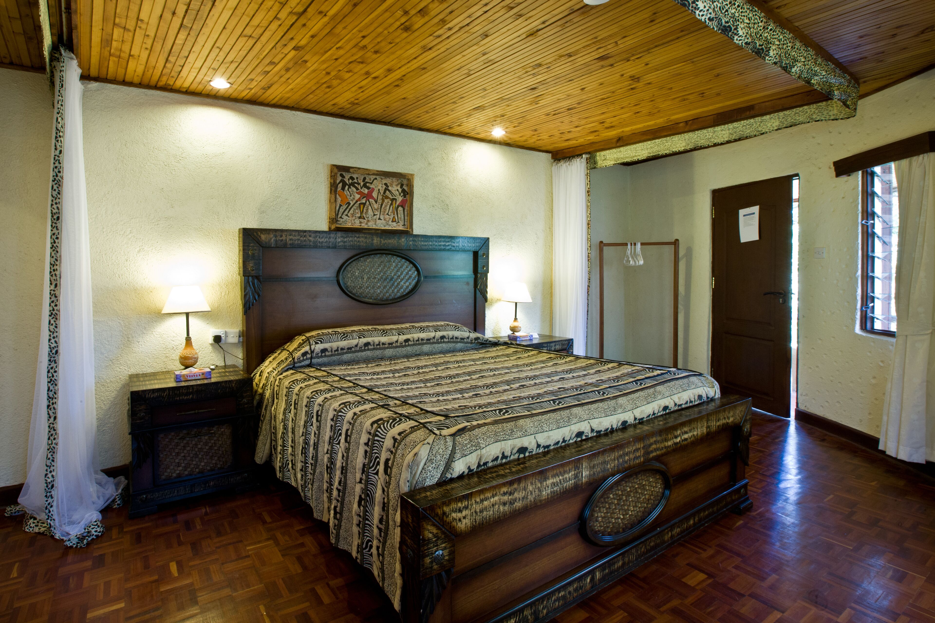 Een traditioneel houten bed met een gebedrukte sprei, twee nachtlampjes en een kunstwerk in een lijst boven het hoofdeinde.