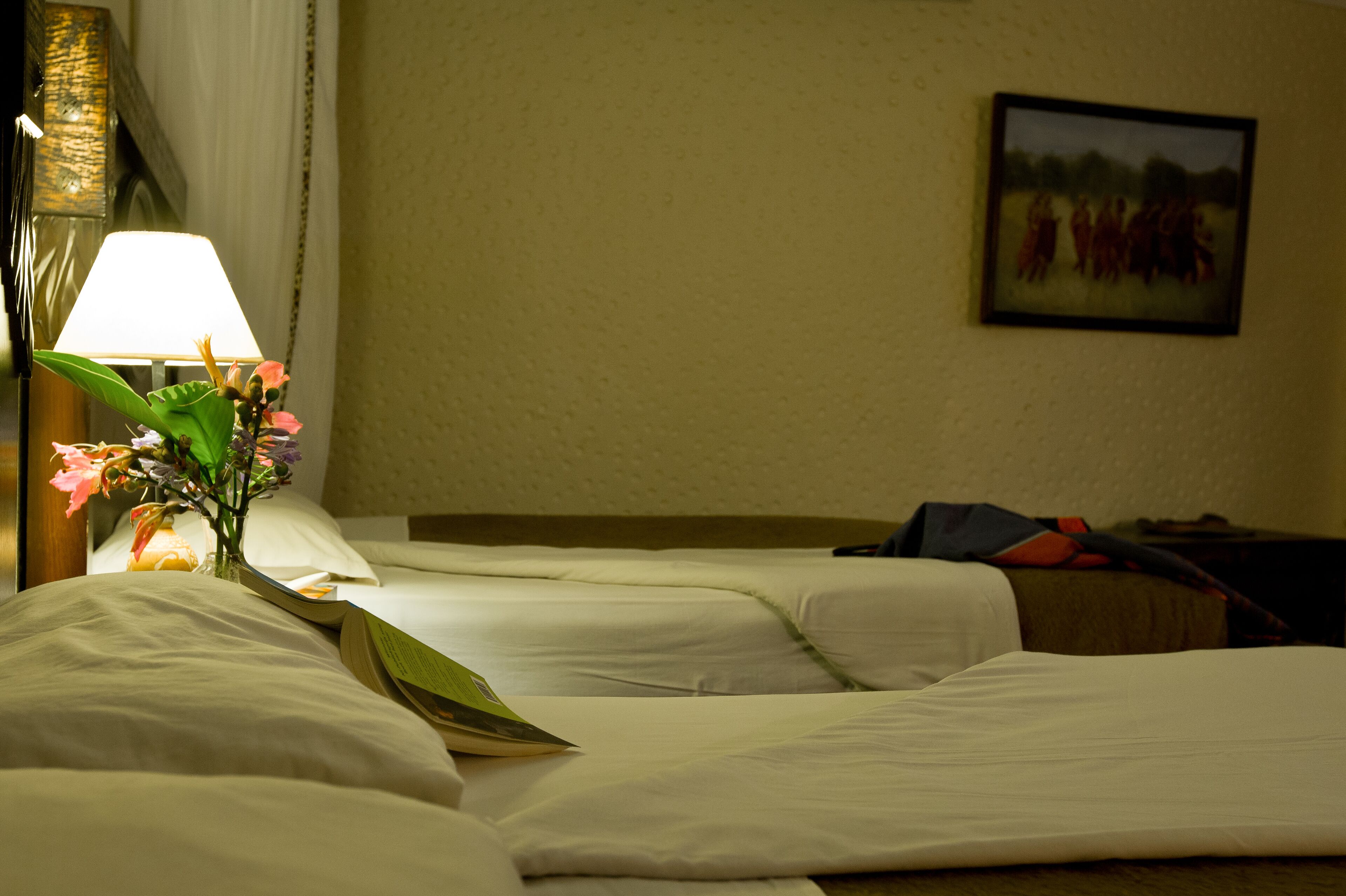 A hotel room with two beds, a bedside lamp, a framed picture on the wall, and a small table with a vase of flowers.