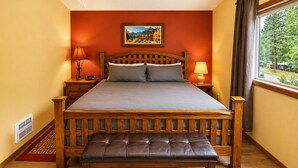 A neatly arranged bedroom with a wooden bed, two bedside tables, a bench, a window with curtains, and a painting on the wall.