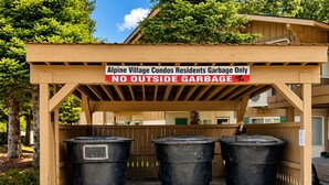 A wooden structure with a sign indicating it is for Alpine Village Condos residents garbage only, with three black trash bins underneath.