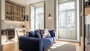 A modern living room with a dark blue sofa, a plaid pillow, and a small round table. There is a kitchen area with a built-in oven and a window with a view of the city. - Líbere Porto Laranjais (Porto)