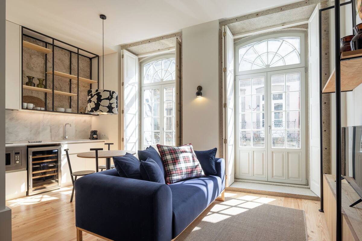 A modern living room with a dark blue sofa, a plaid pillow, and a small round table. There is a kitchen area with a built-in oven and a window with a view of the city.