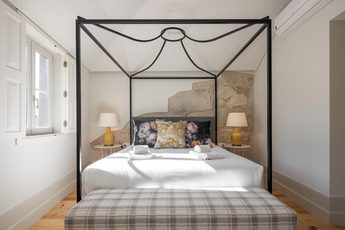 A four-poster bed with a white bedspread and a plaid throw, flanked by two bedside tables with lamps, in a room with a stone accent wall and a window with white shutters.