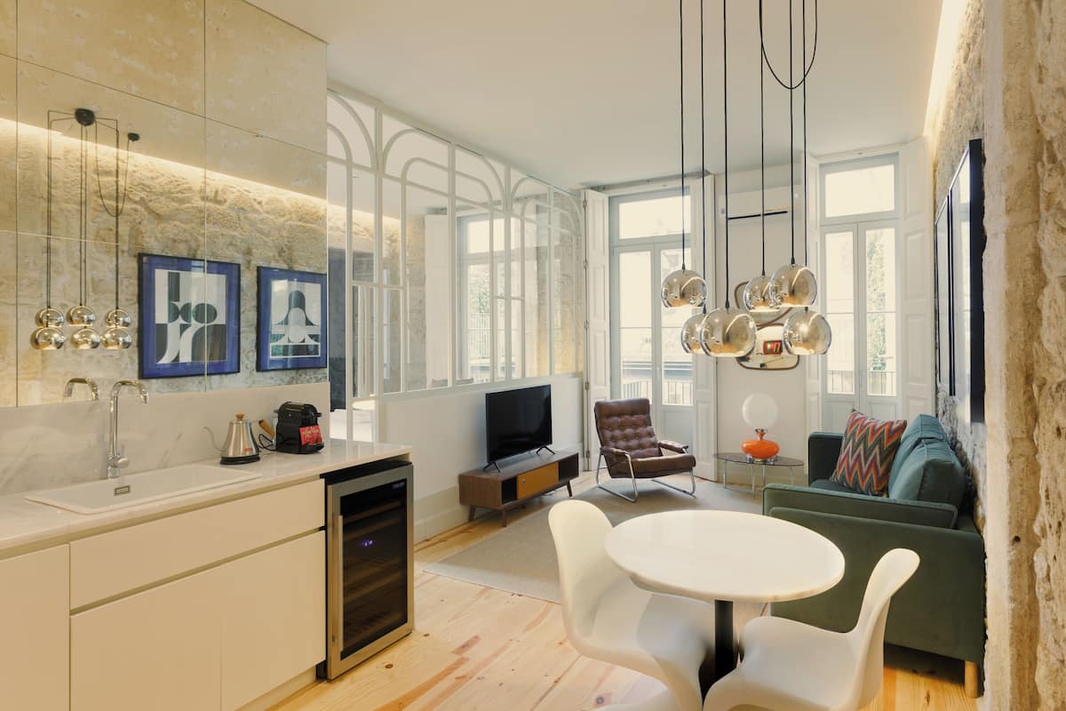 A modern kitchen with a built-in oven, a sink, and a television. There is a dining area with a white table and chairs, and a green sofa with a colorful pillow.