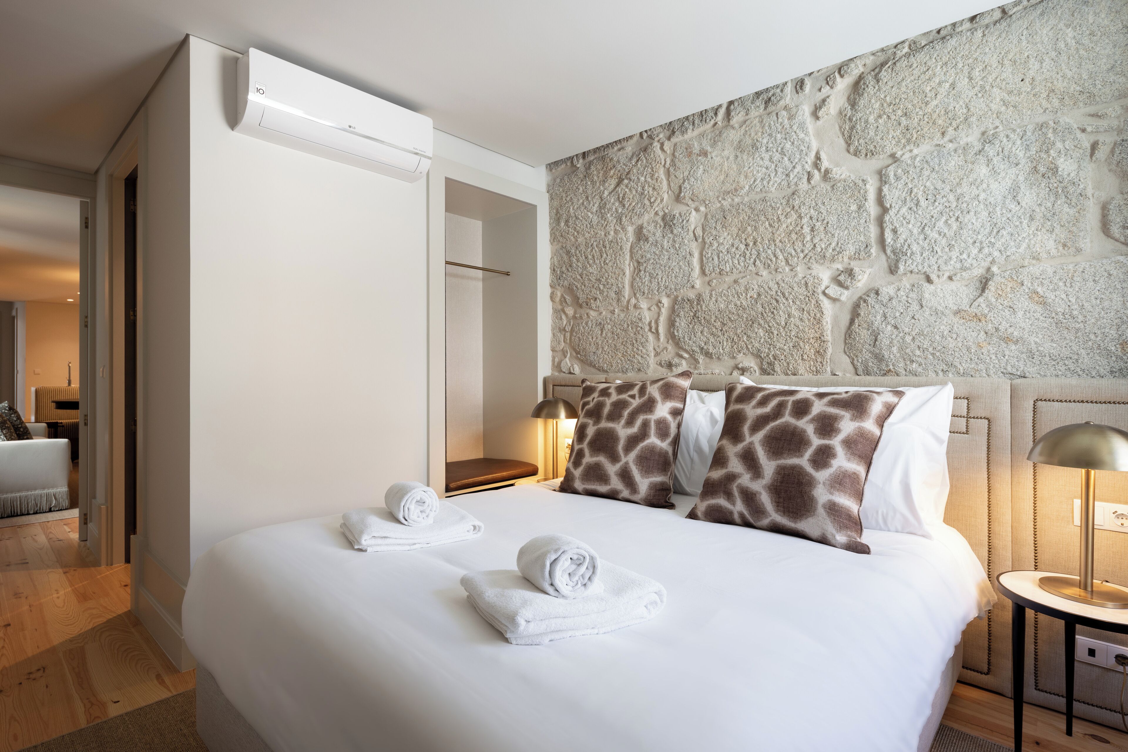 A hotel room with a large bed, a stone wall, a bedside table with a lamp, and a bathroom visible through an open door.