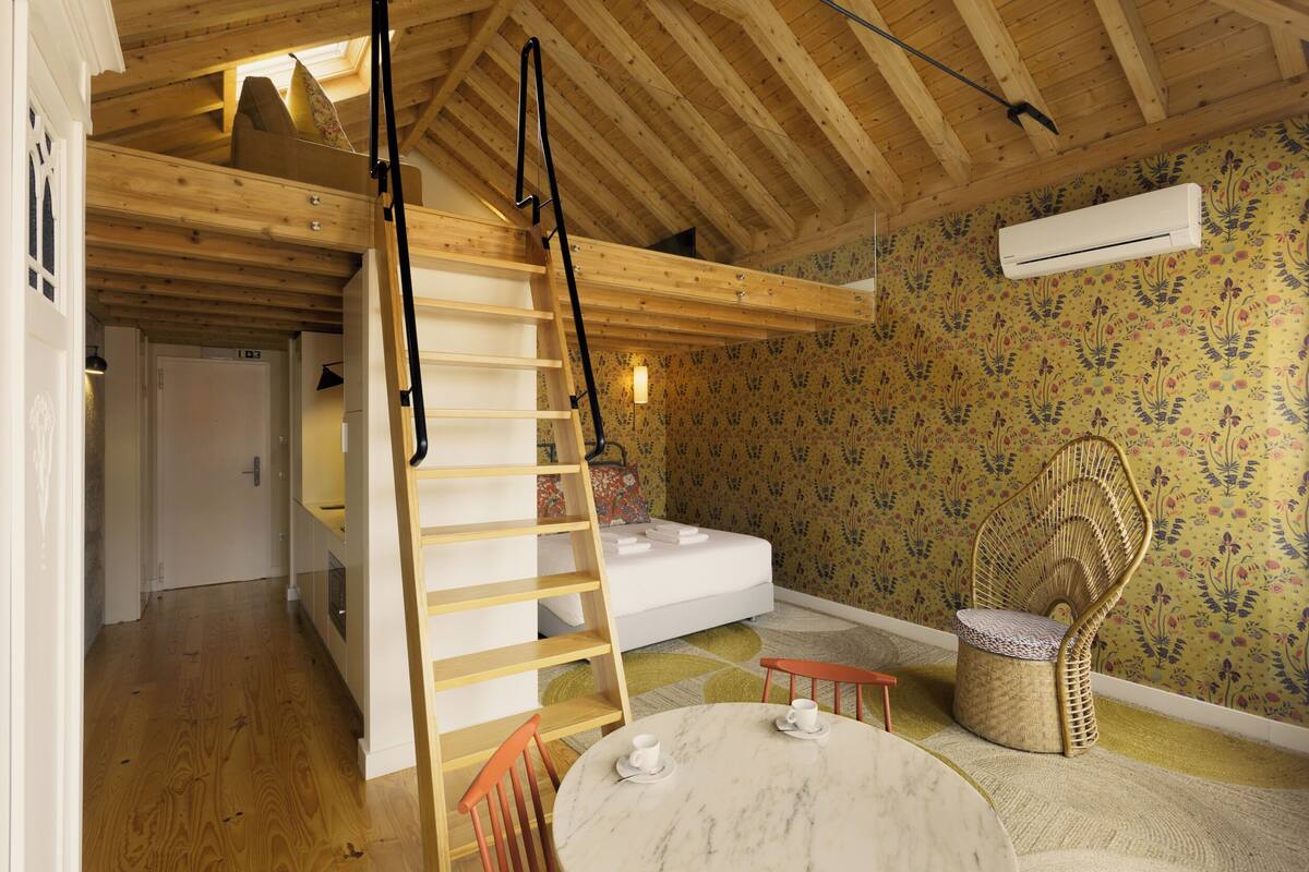 A room with a wooden ceiling, a bunk bed, a small table with two chairs, a wicker chair, and a patterned wallpaper.