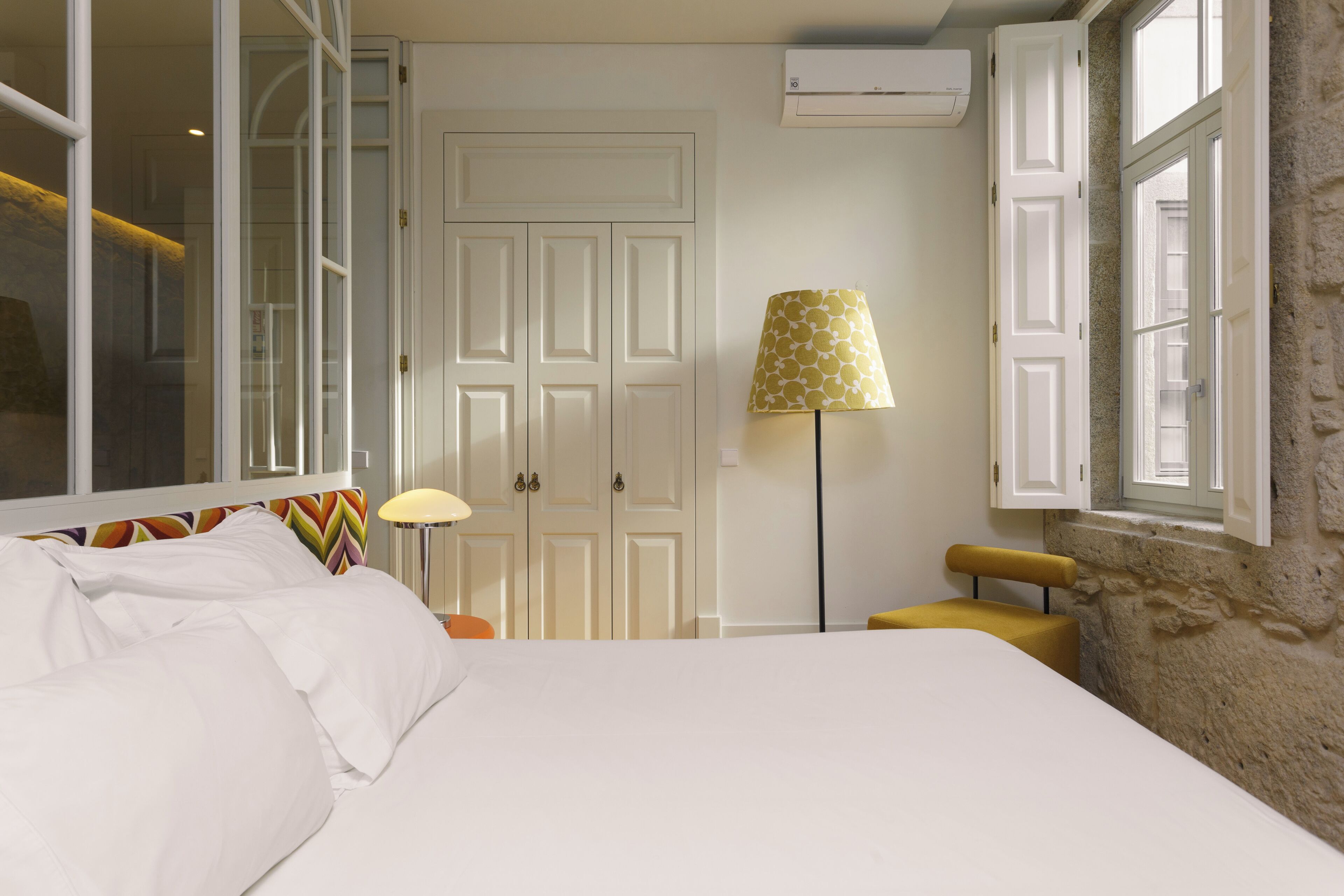 A hotel room with a bed, a lamp, a chair, a wardrobe, and a window.