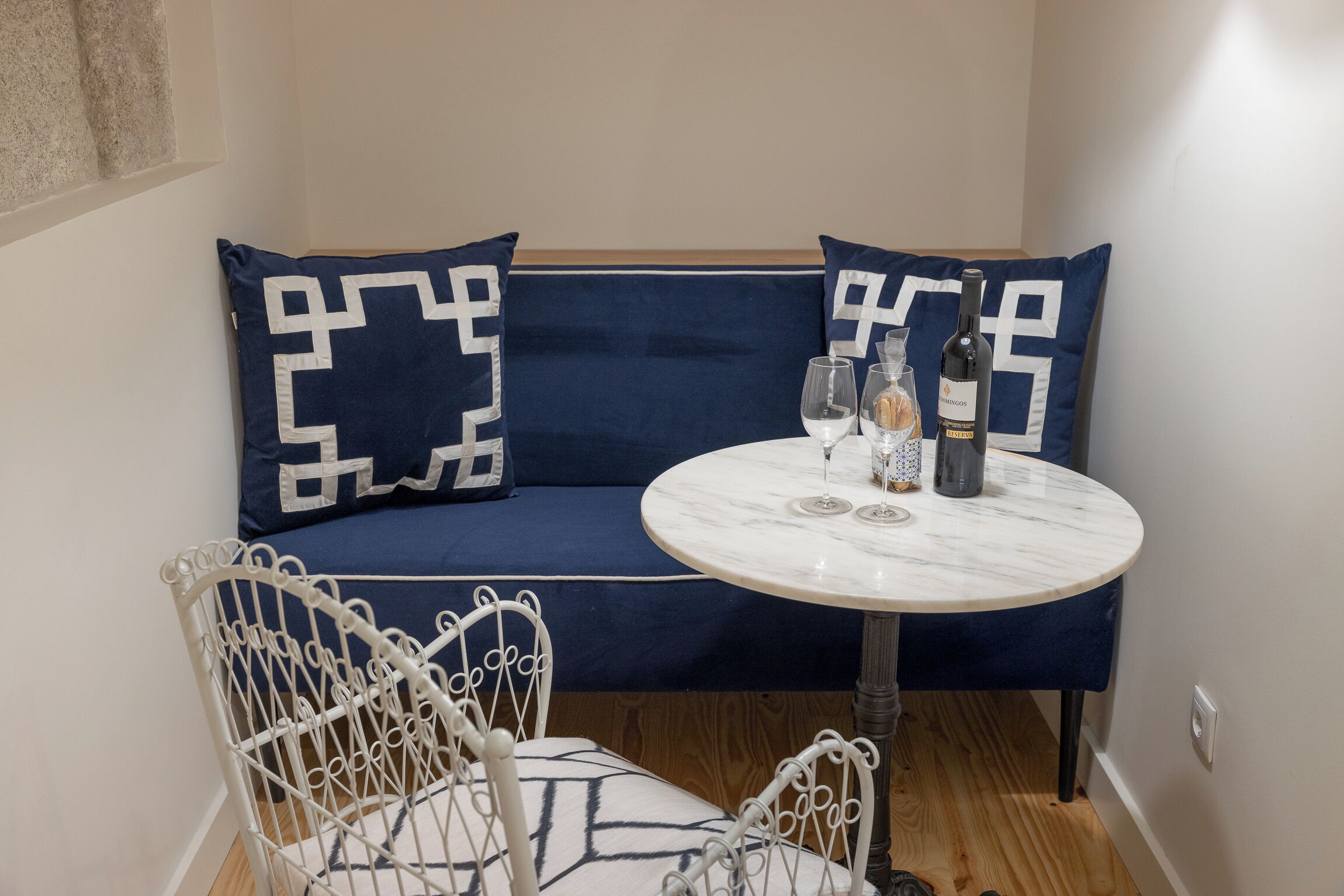 A corner seating area with a blue sofa and matching pillows, a round marble table with a bottle and glasses, and white metal chairs.