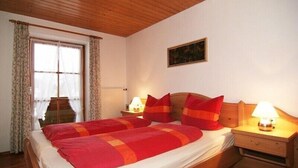 A double bed with red and orange bedding, two bedside tables with lamps, a window with curtains, and a painting on the wall. - Königssee, vacation apartments Klausner (Schönau am Königssee)