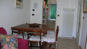 Een eetgedeelte met een houten tafel en stoelen, een bloemenkussen op een bank, en uitzicht op een tuin door een boog.