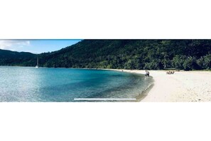 Une plage avec un bateau sur l’eau, un rivage sablonneux et une colline boisée en arrière-plan.