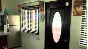 Une petite cuisine équipée d’un réfrigérateur, d’une fenêtre avec des stores, d’une horloge et d’une porte donnant sur l’extérieur.
