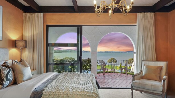 A bedroom with a large bed, a chair, and a view of the sunset through a glass door.