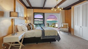 A bedroom with a bed, bedside table, lamp, chair, and a view of the sea through a window.