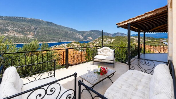 A balcony with a white cushioned chair, a small table with a bottle and flowers, and a view of a coastal landscape with mountains.