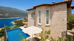 A stone building with a pool, a patio with a table and chairs, and a view of the sea and mountains.