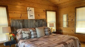 Ein Schlafzimmer einer Holzhütte mit einem Bett, Karo-Kissen, einem Nachttisch, einer Lampe und einem Schild an der Wand.