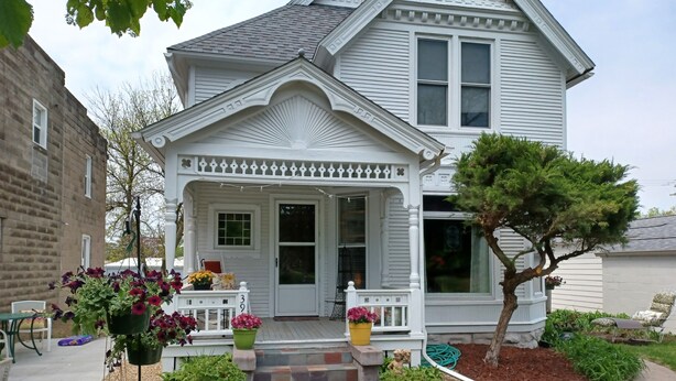 A two-story house with a white porch, a front door, and a small garden area with potted plants and a tree.