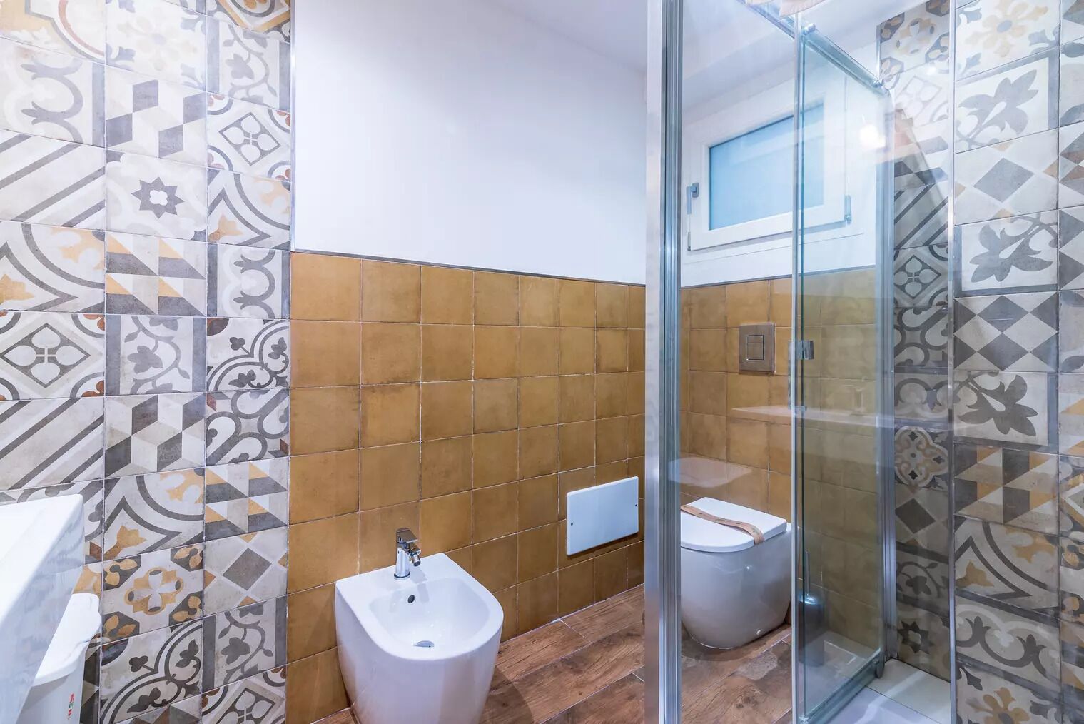 A bathroom with patterned tiles, a bidet, a toilet, and a glass shower enclosure.