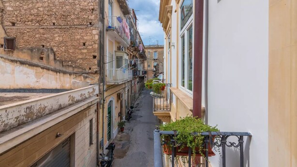 Una calle estrecha flanqueada por edificios, con balcones adornados con plantas en macetas.