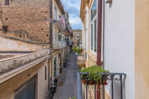 A narrow street with buildings on either side, featuring balconies with potted plants.