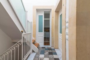 A narrow hallway with a tiled floor, a staircase with white railings, and a door leading to a room with a tiled backsplash.