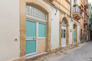 A narrow street with buildings featuring large windows and doors, some with shutters.