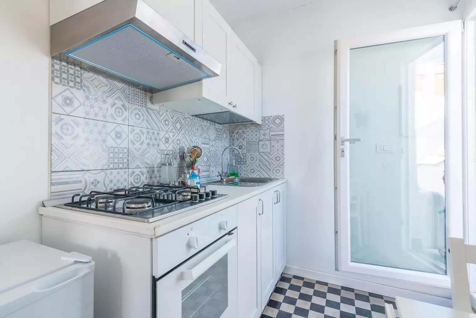 A compact kitchen with a gas stove, white cabinets, patterned backsplash, and a checkered floor.