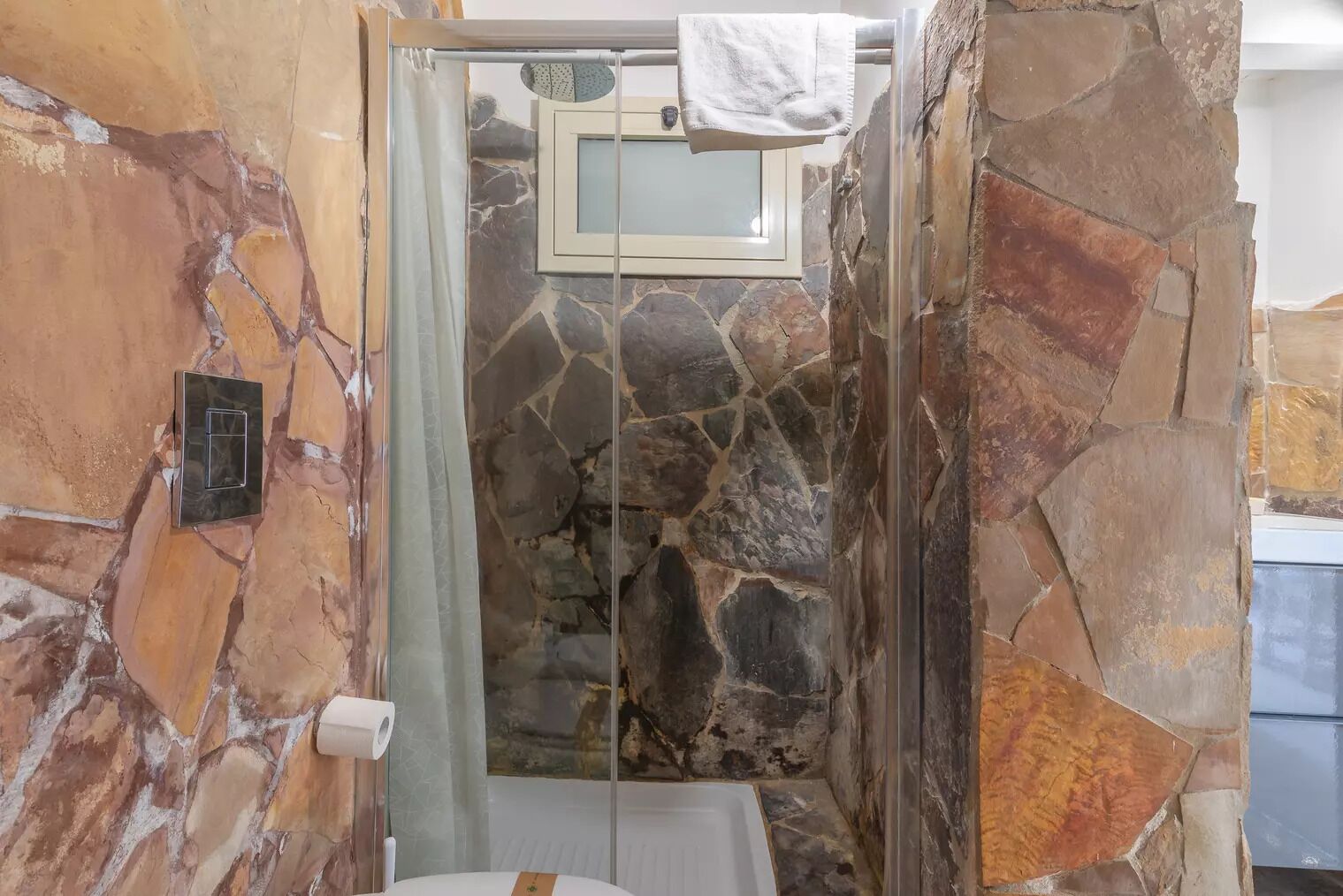 A bathroom with a stone wall, a glass shower enclosure, a towel hanging on a rack, and a toilet paper holder.