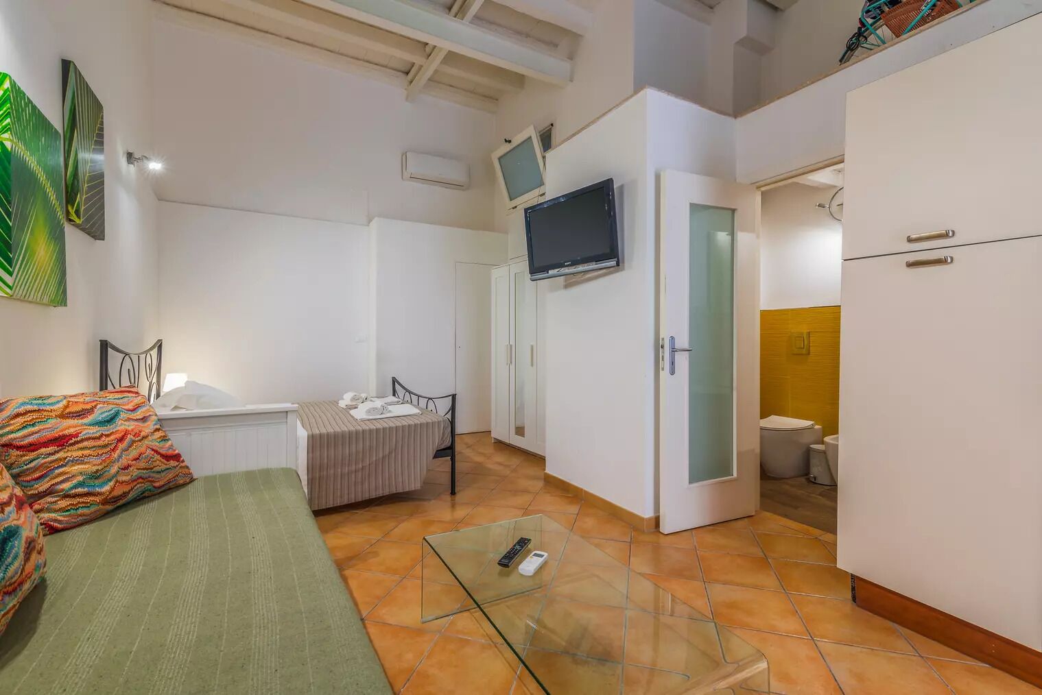A compact room with a bed, a small table, a TV mounted on the wall, and a wardrobe.