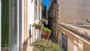 Una calle urbana estrecha flanqueada por edificios, con un pequeño balcón y plantas en macetas, y un cielo azul despejado.