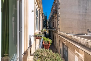 A narrow urban street with buildings on either side, a small balcony with potted plants, and a clear blue sky.