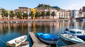 Una costa con barcos amarrados a un muelle de madera, un edificio histórico con una fortaleza en una colina y palmeras bordeando el agua.