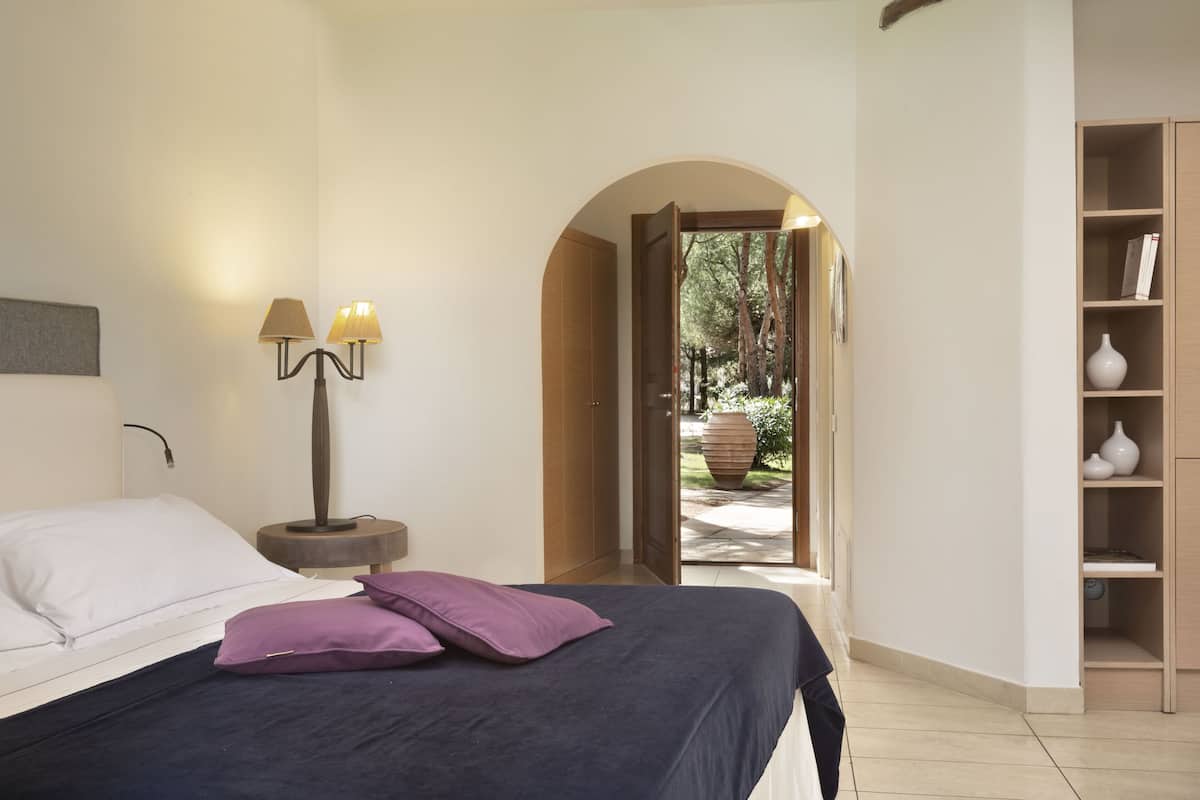Une chambre à coucher avec un lit, une table de chevet avec des lampes, une bibliothèque et une porte en arc donnant sur un jardin.