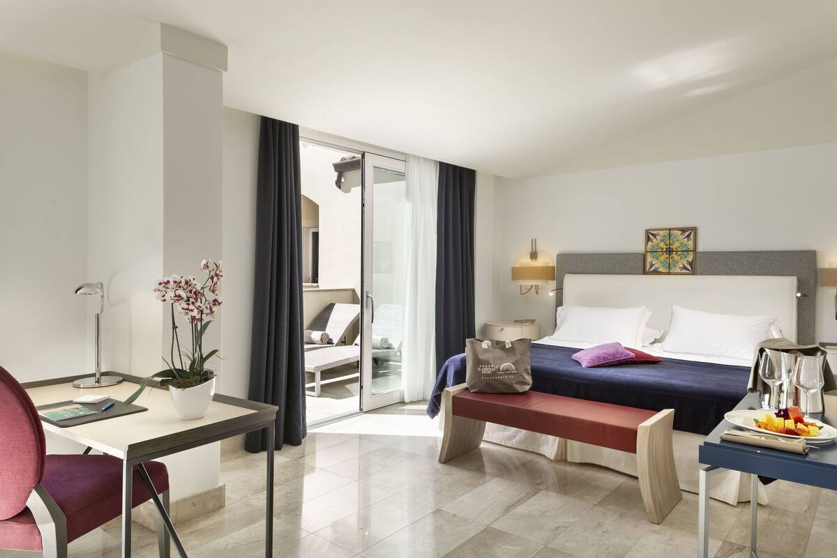 Une chambre d’hôtel avec un lit, un bureau avec une chaise, un banc, une table à manger avec une assiette de fruits et une plante en pot.