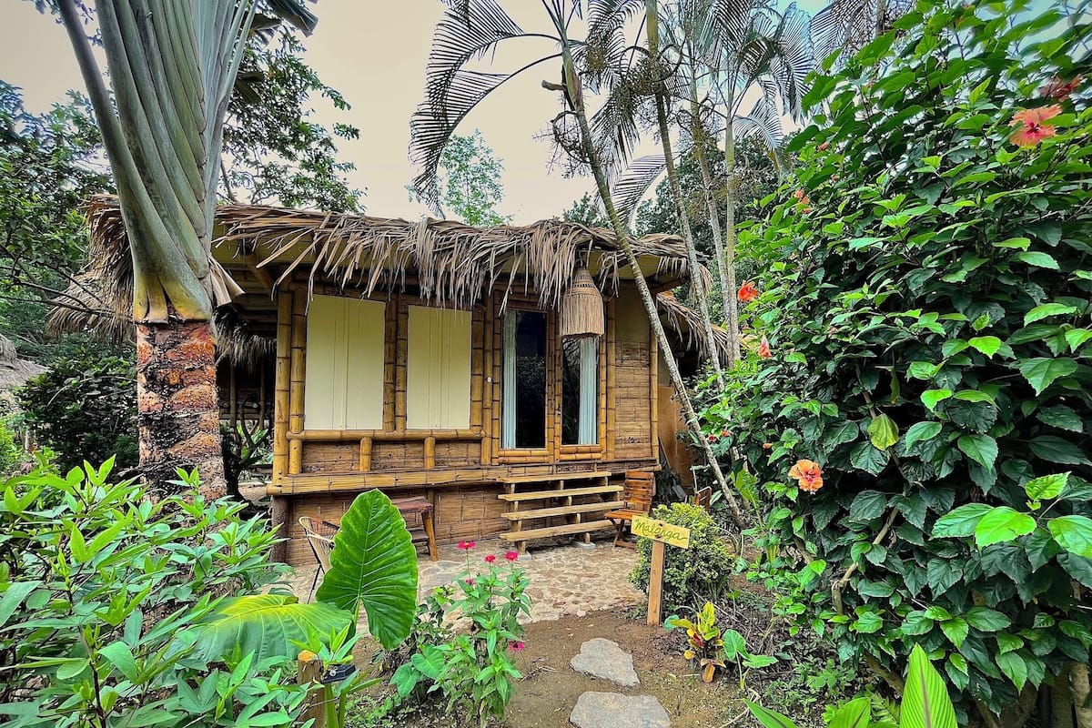 A rustic bamboo hut with a thatched roof, surrounded by lush greenery and a stone pathway leading to the entrance.