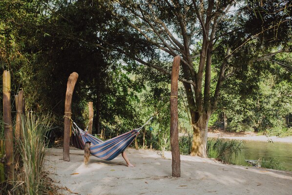 A hammock tied between trees in a natural setting with a sandy path and a body of water nearby. - YAY Sustainable (Santa Marta)