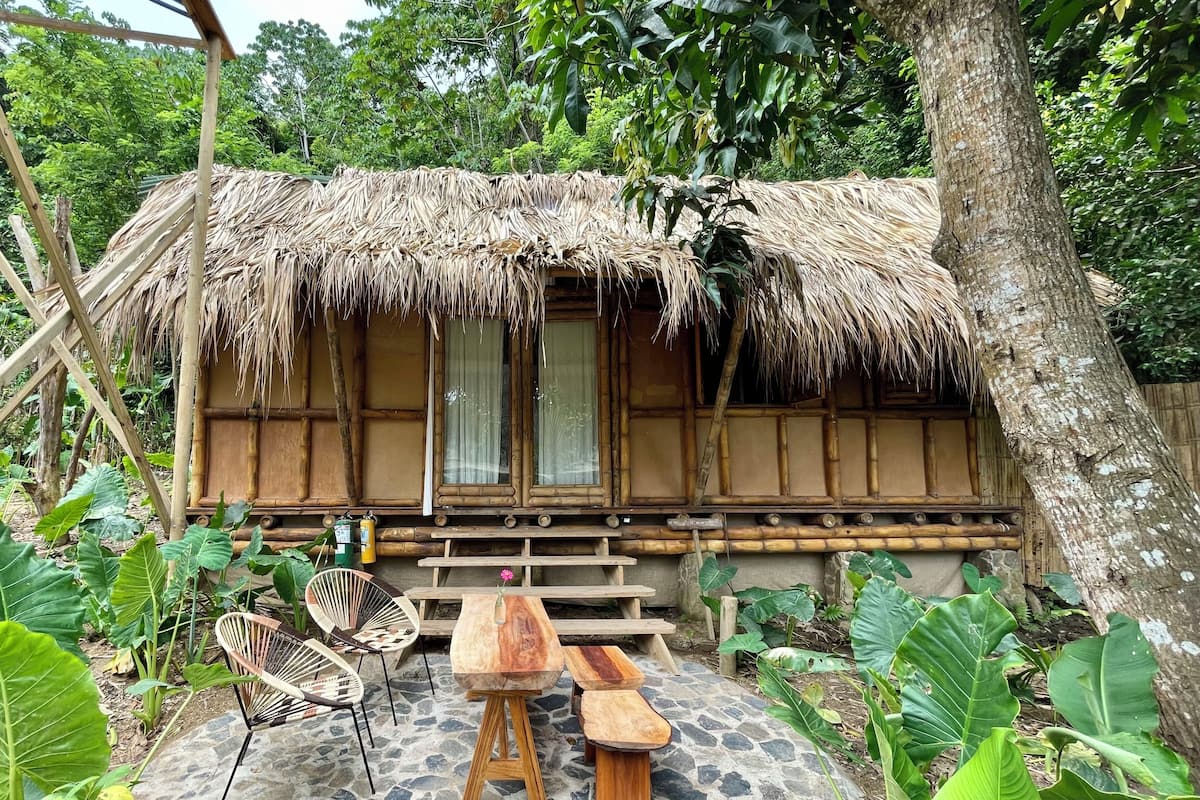 A rustic thatched-roof hut with a stone-paved patio, wooden furniture, and lush greenery.
