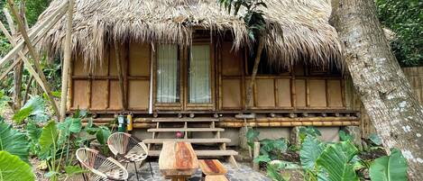A rustic thatched-roof hut with a stone-paved patio, wooden furniture, and lush greenery.